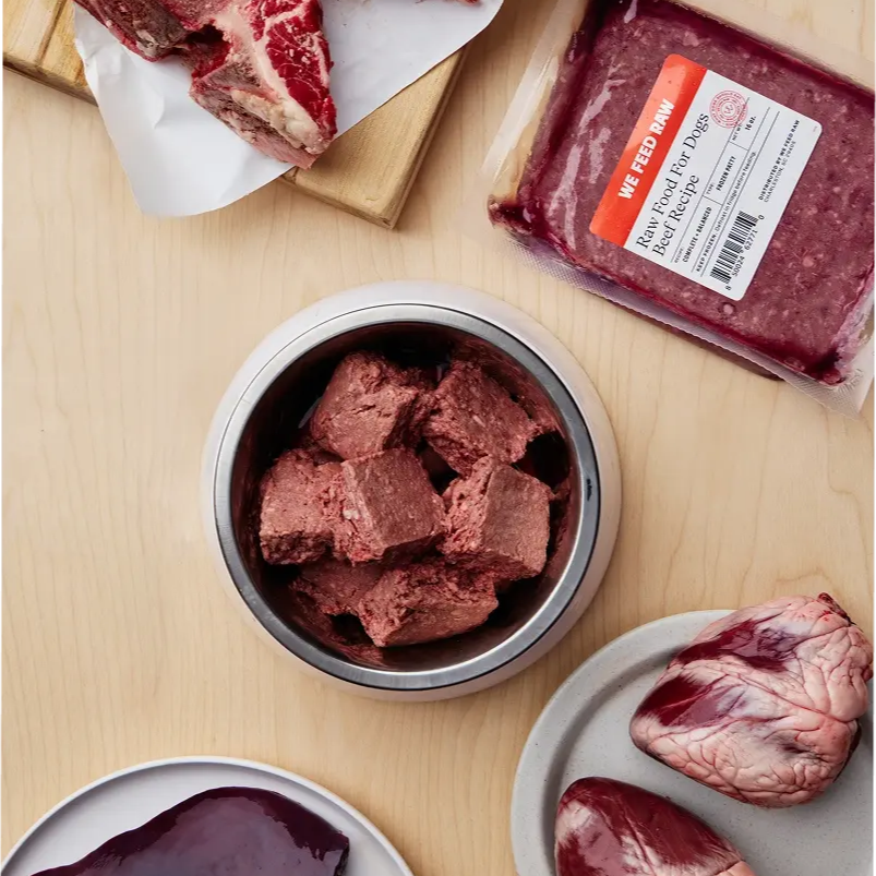 Various cuts of raw meat and prepared raw dog food displayed on a wooden cutting board with butcher knife and kitchen towels. Includes raw muscle meat, organs, and ground meat in a metal container.