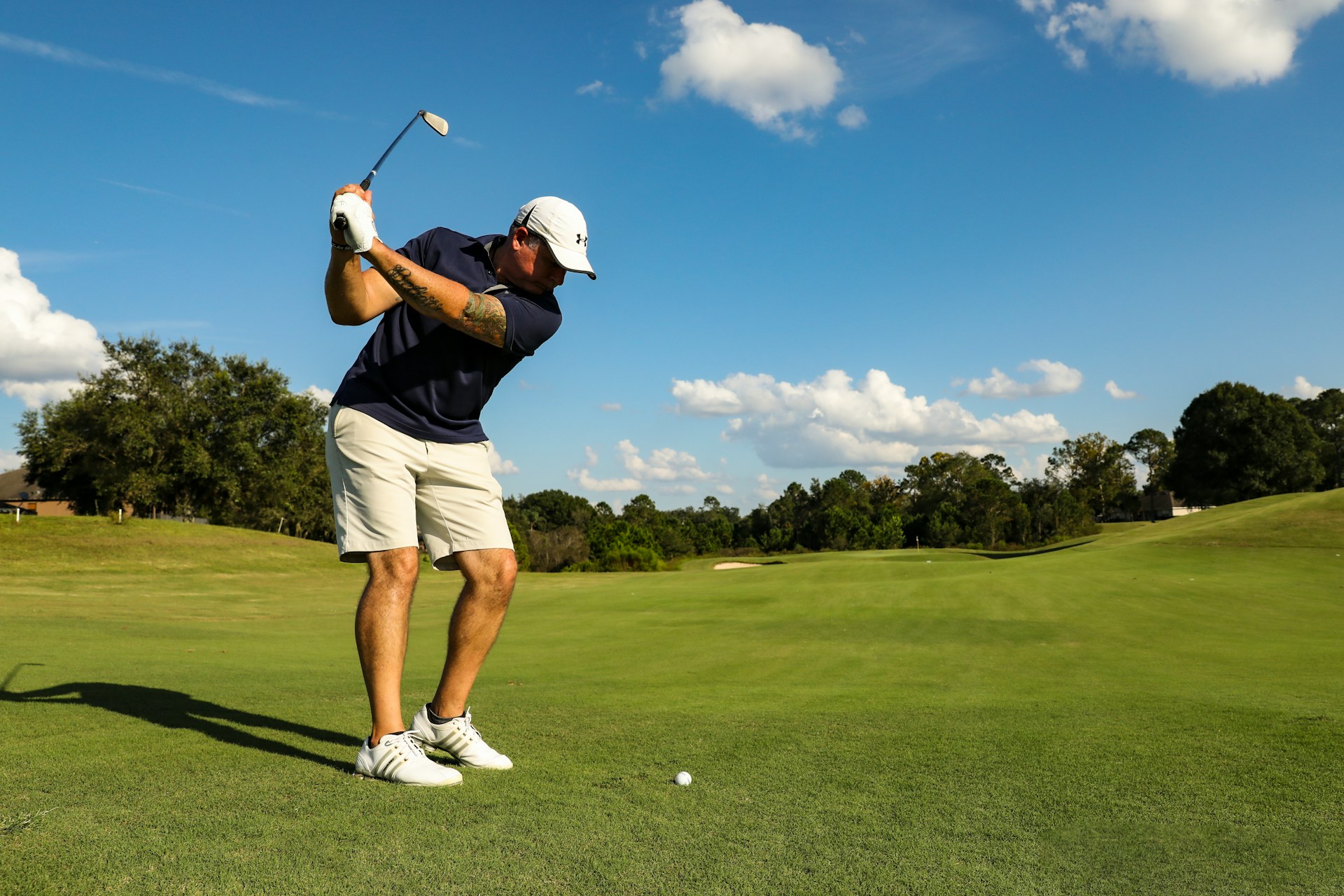 Golfer in mid-swing on a sunny golf course with trees and blue sky in the background.