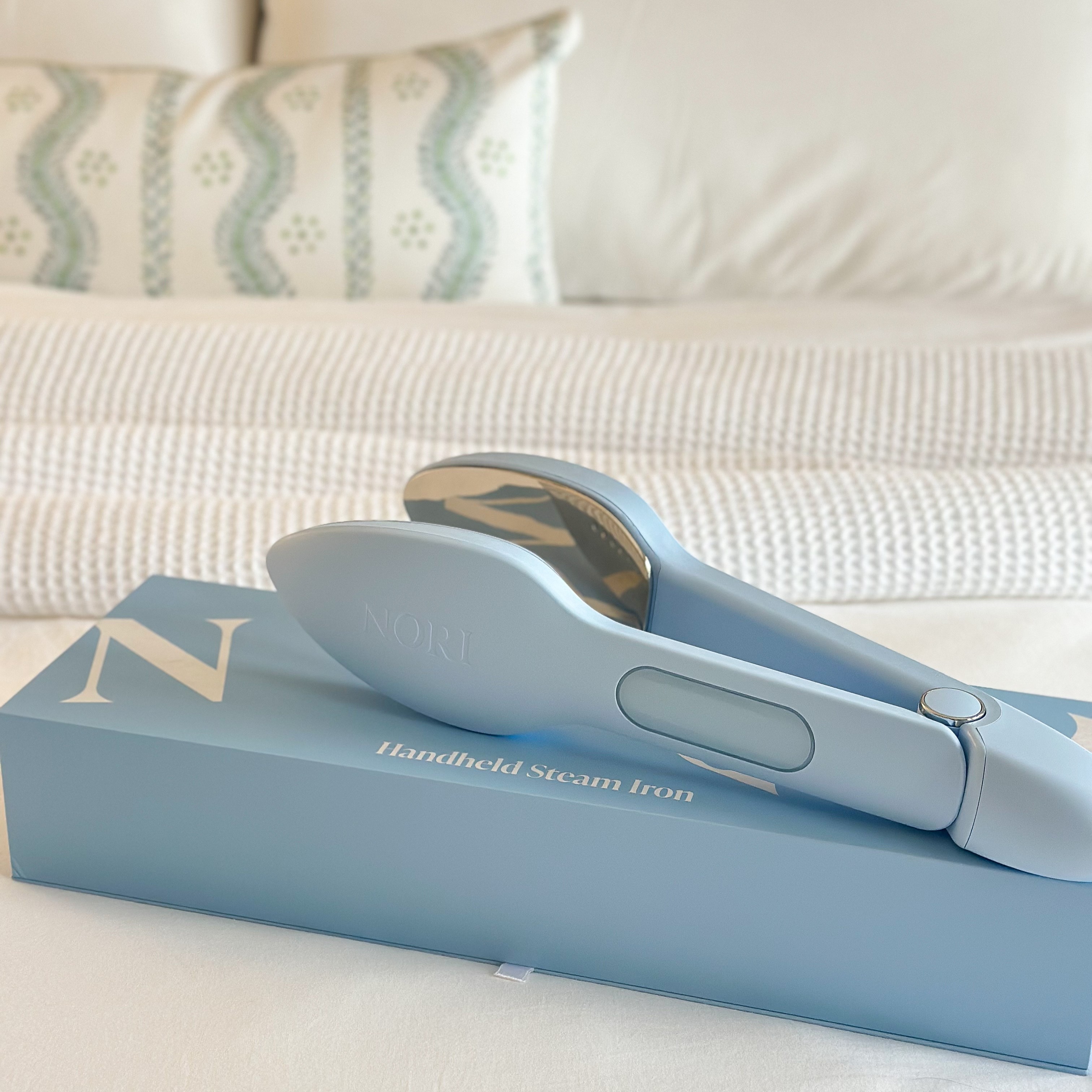 The Nori Press in blue resting on its box on top of a white bed
