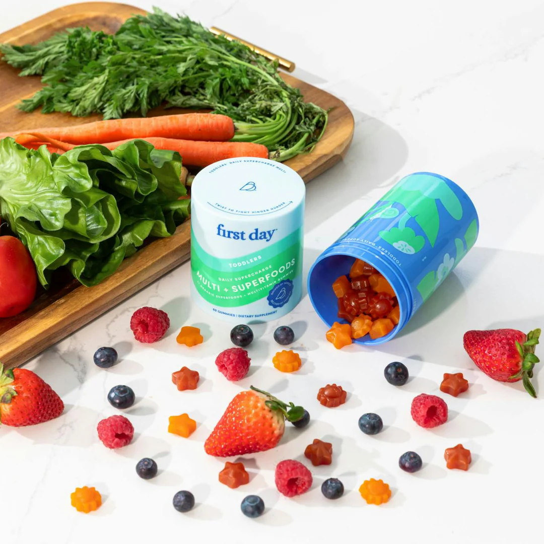 First Day multivitamins for kids are displayed with fresh fruits and vegetables.