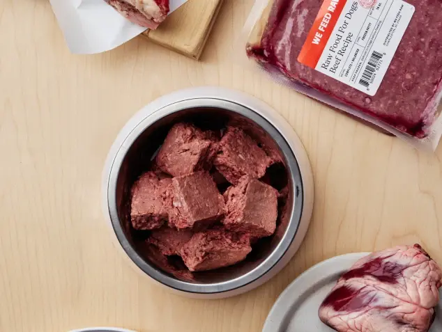 A bowl of We Feed Raw dog food cubes is surrounded by packaged raw meat on a wooden table