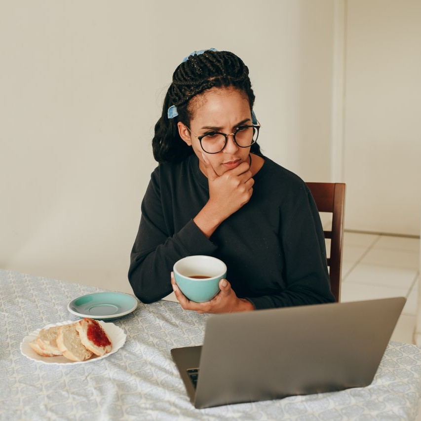 Woman with glasses holds a cup and looks at a laptop with a thoughtful expression