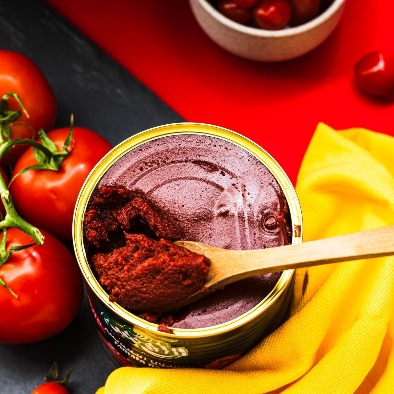 A can of tomato paste with a wooden spoon surrounded by fresh tomatoes