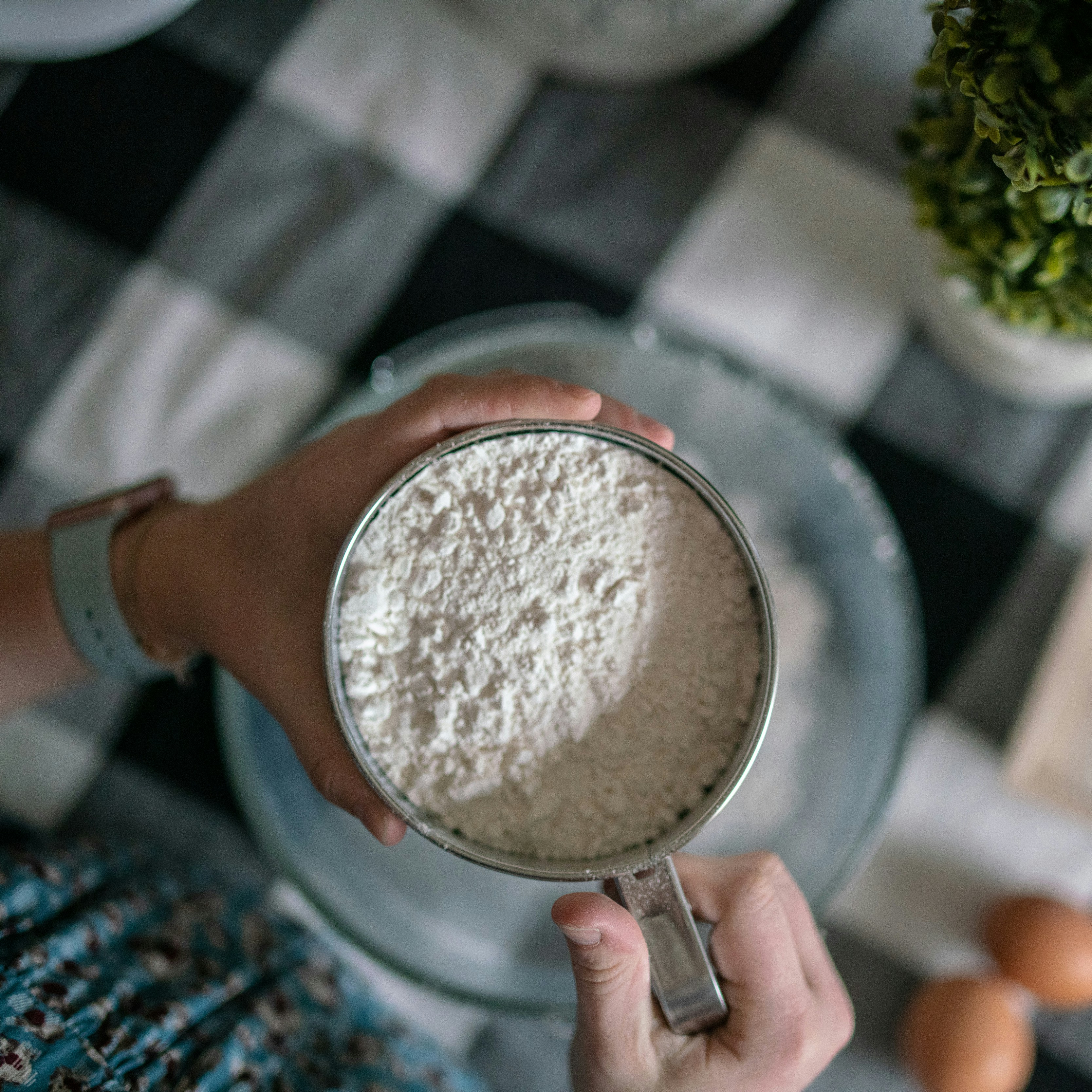 Person holds a measuring cup filled with flour over a bowl on a checkered tablecloth