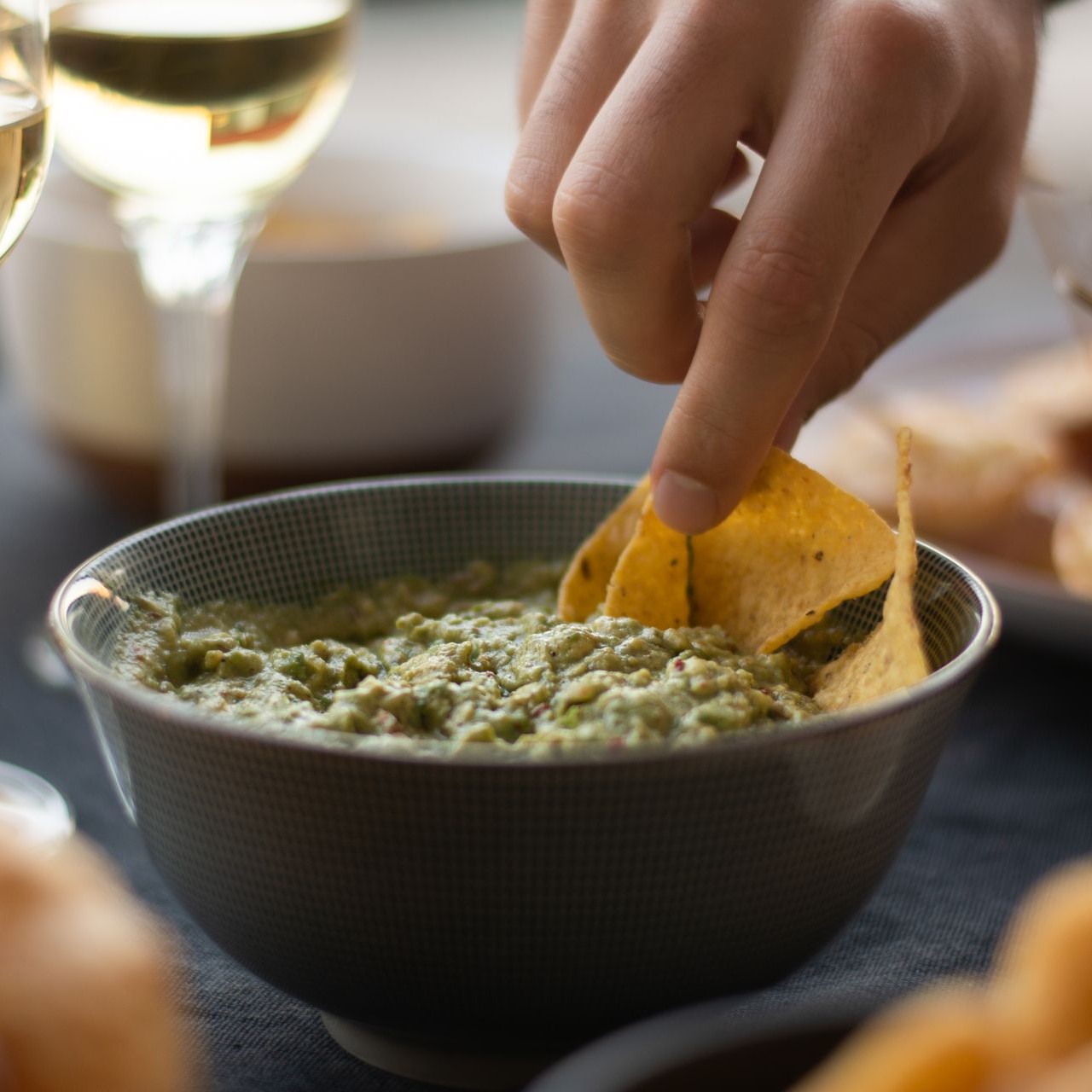A hand dips a tortilla chip into a bowl of guacamole at a party