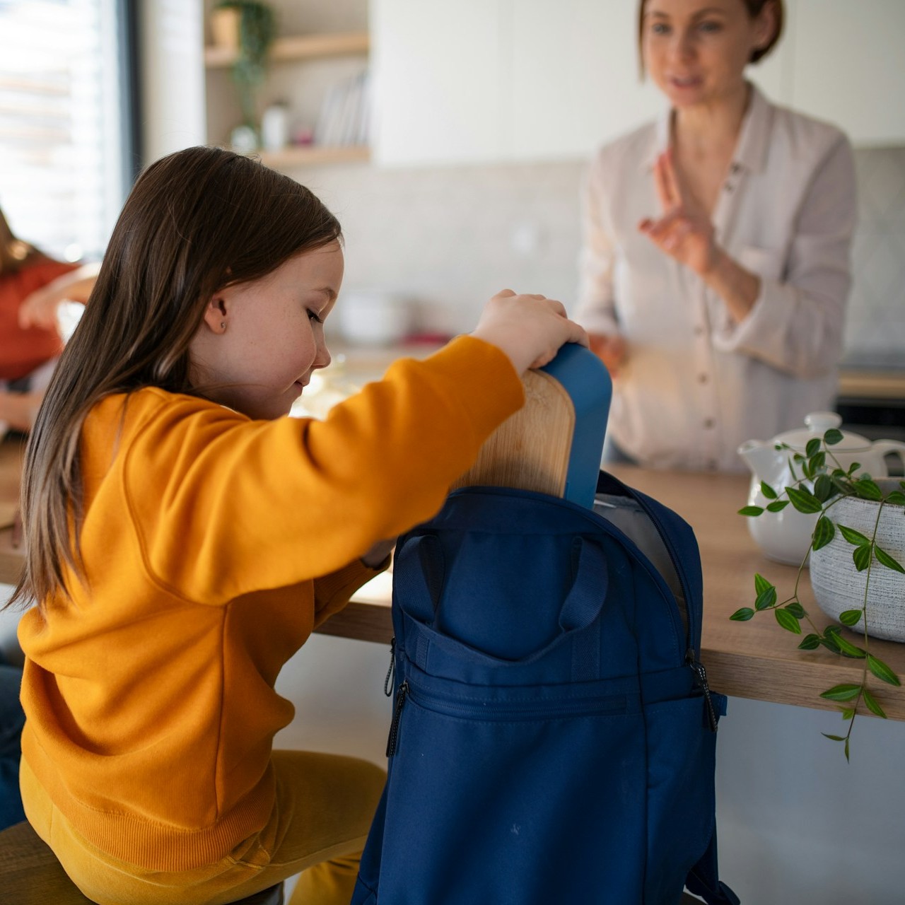 Child Unpacking Backpack in Kitchen Child unpacks a backpack at a kitchen counter while her mom talks nearby