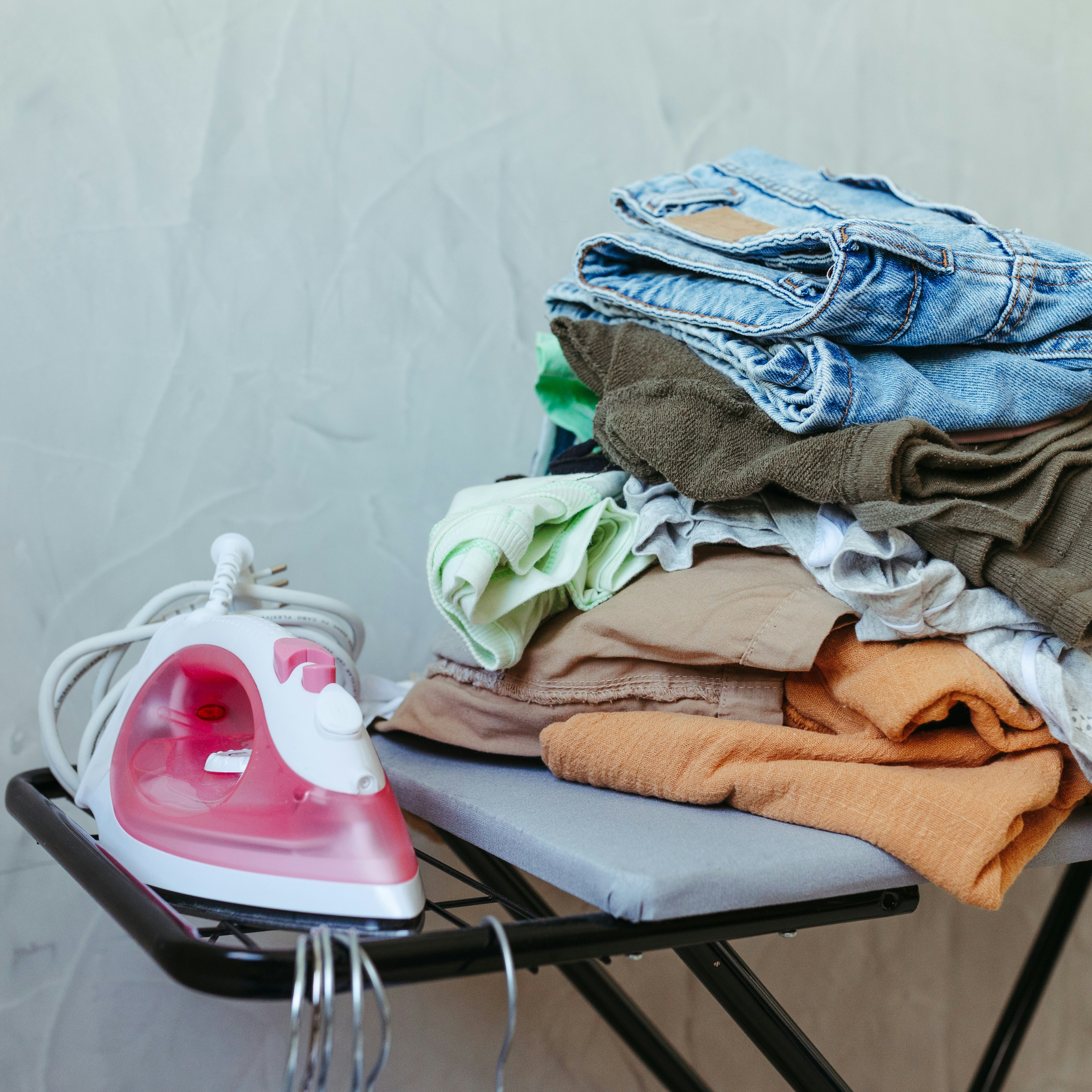 A small ironing board holds a stack of folded clothes and a pink iron in a compact space