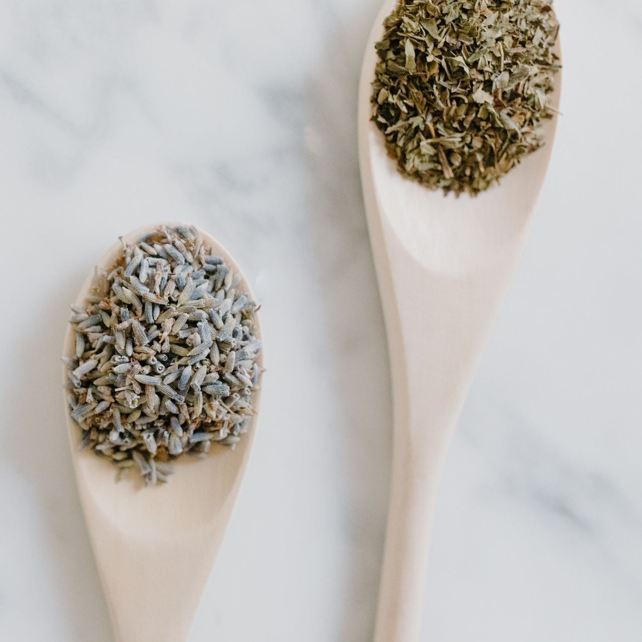 Two wooden spoons hold dried herbs on a marble surface