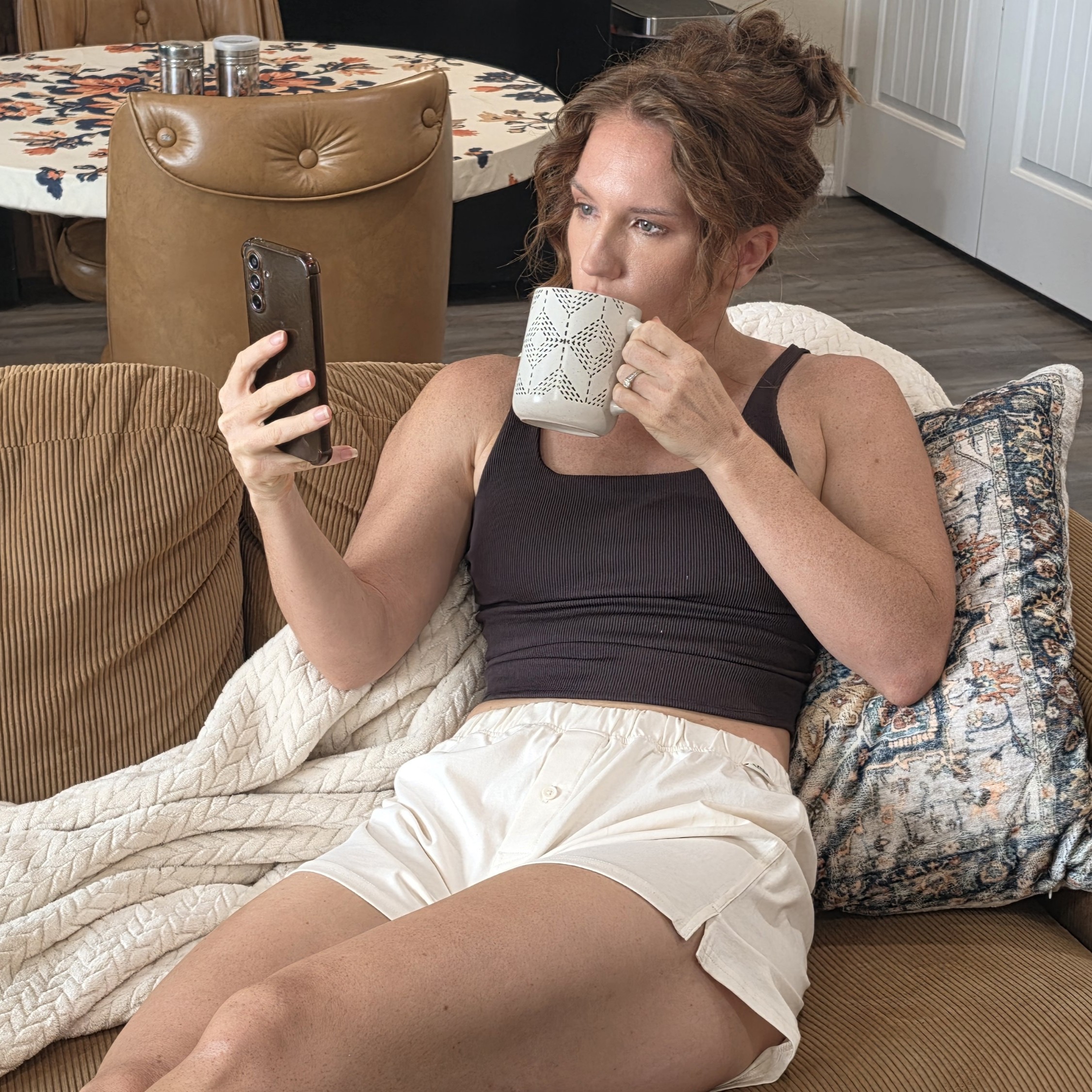Researching on the Couch with Coffee Abby lounges on a couch, drinking from a mug and researching on her phone while wearing NADS Ball Natural™ boxers